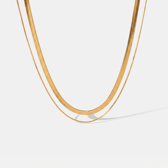 A layered gold-tone necklace of two flat herringbone chains, on a plain background