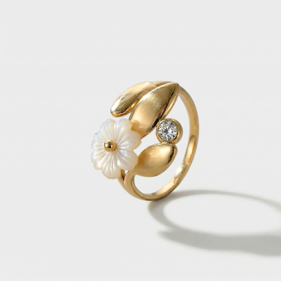 A gold-tone open ring with a white blossom among leaves and a crystal, on a plain background